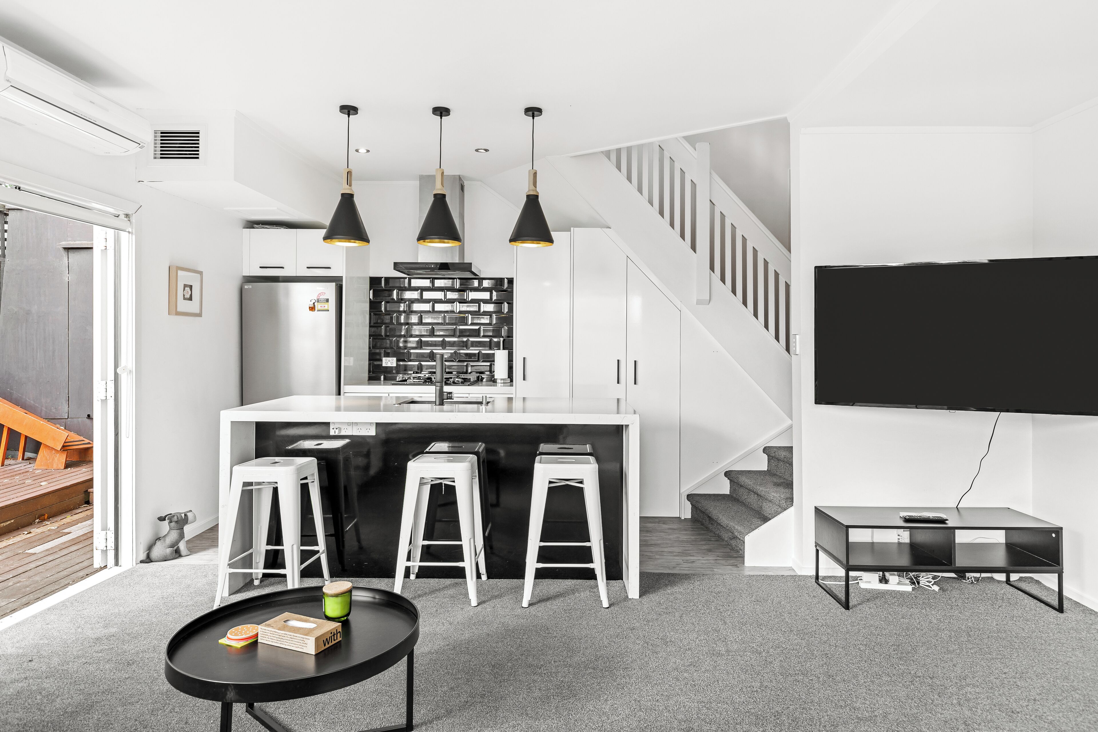 A modern kitchen with white cabinets, a stainless steel refrigerator, and a black backsplash. There are three white stools and a black coffee table with a remote control. A staircase with a white railing leads to another level.