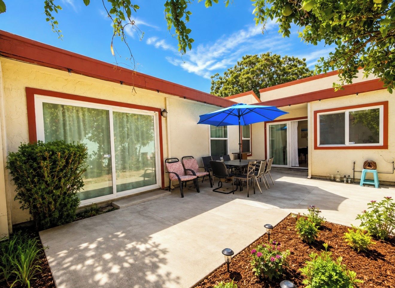 A residential patio with a table and chairs, a blue umbrella, and a small garden area.