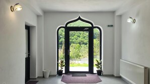 En hall med en stor, välvd dörröppning som leder ut till ett frodigt, grönskande utomhusområde. På golvet, nära dörröppningen, står två krukväxter, och på den högra väggen syns ett vitt element.