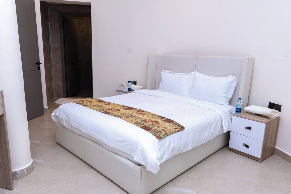 A neatly made bed with a white comforter and a patterned throw, a bedside table with a water bottle, and a nightstand with a phone.
