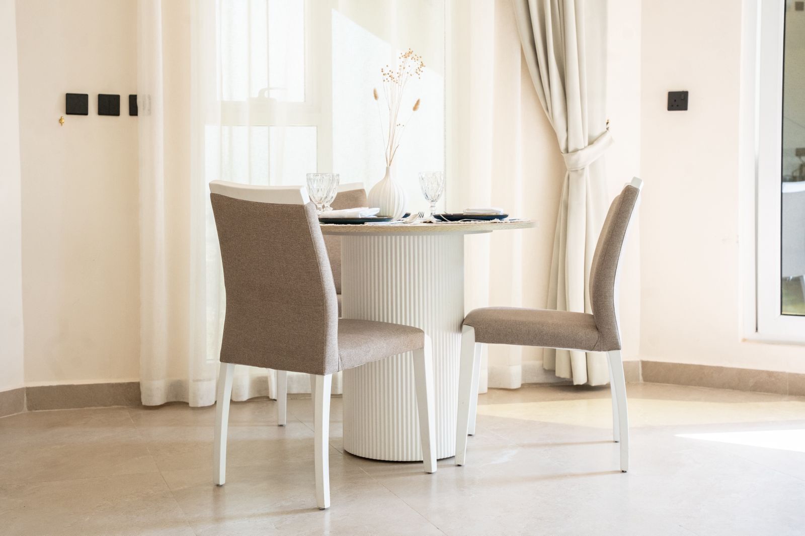 A dining area with a round table, two chairs, and a vase with dried flowers.