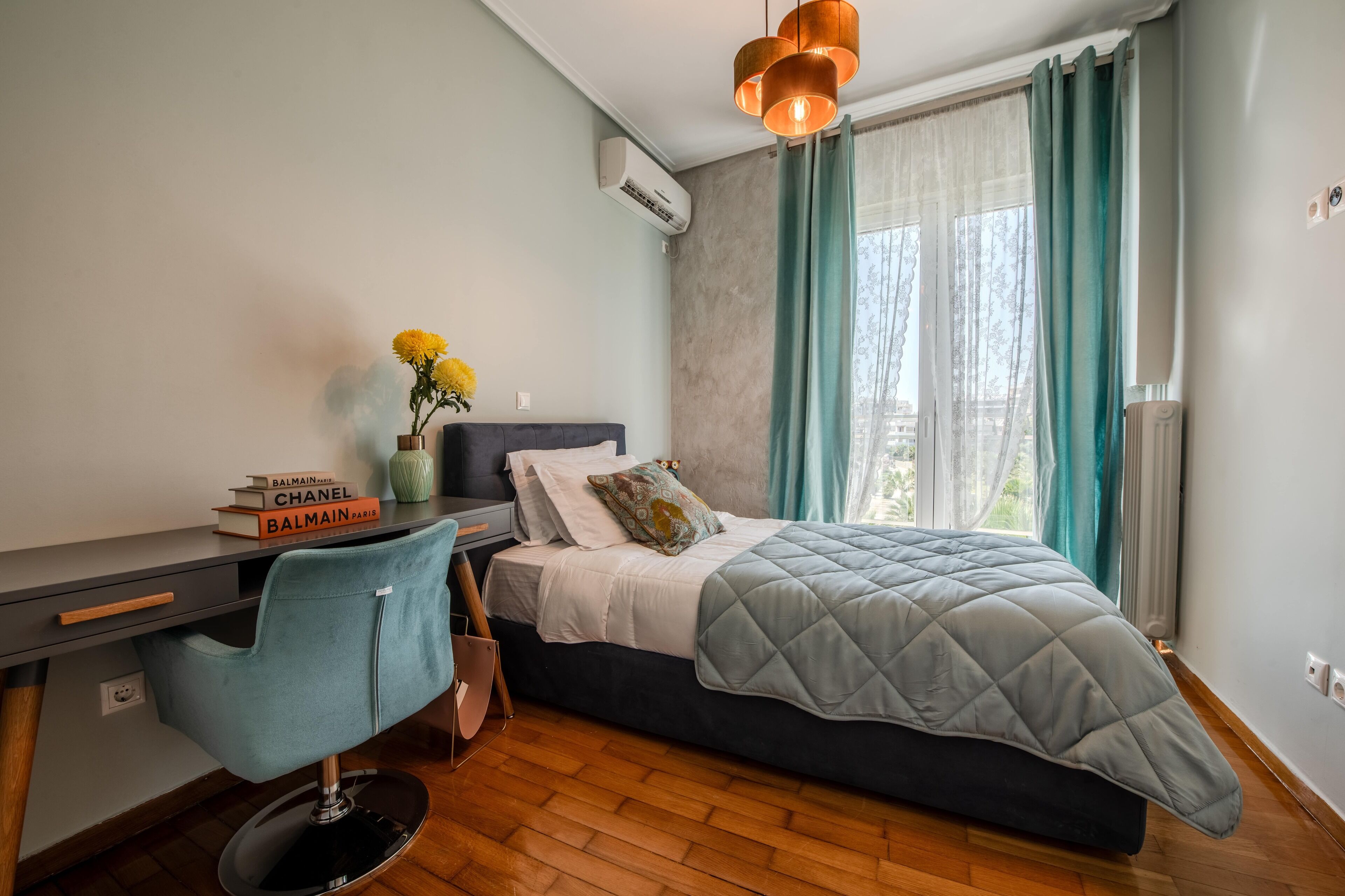 Een slaapkamer met een bed, een bureau en een stoel. Er is een raam met gordijnen, een airconditioner en een vaas met bloemen op het bureau.