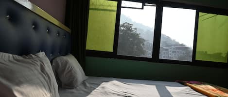 Una cama con cabecera, una ventana que da a edificios y colinas, y una cortina.