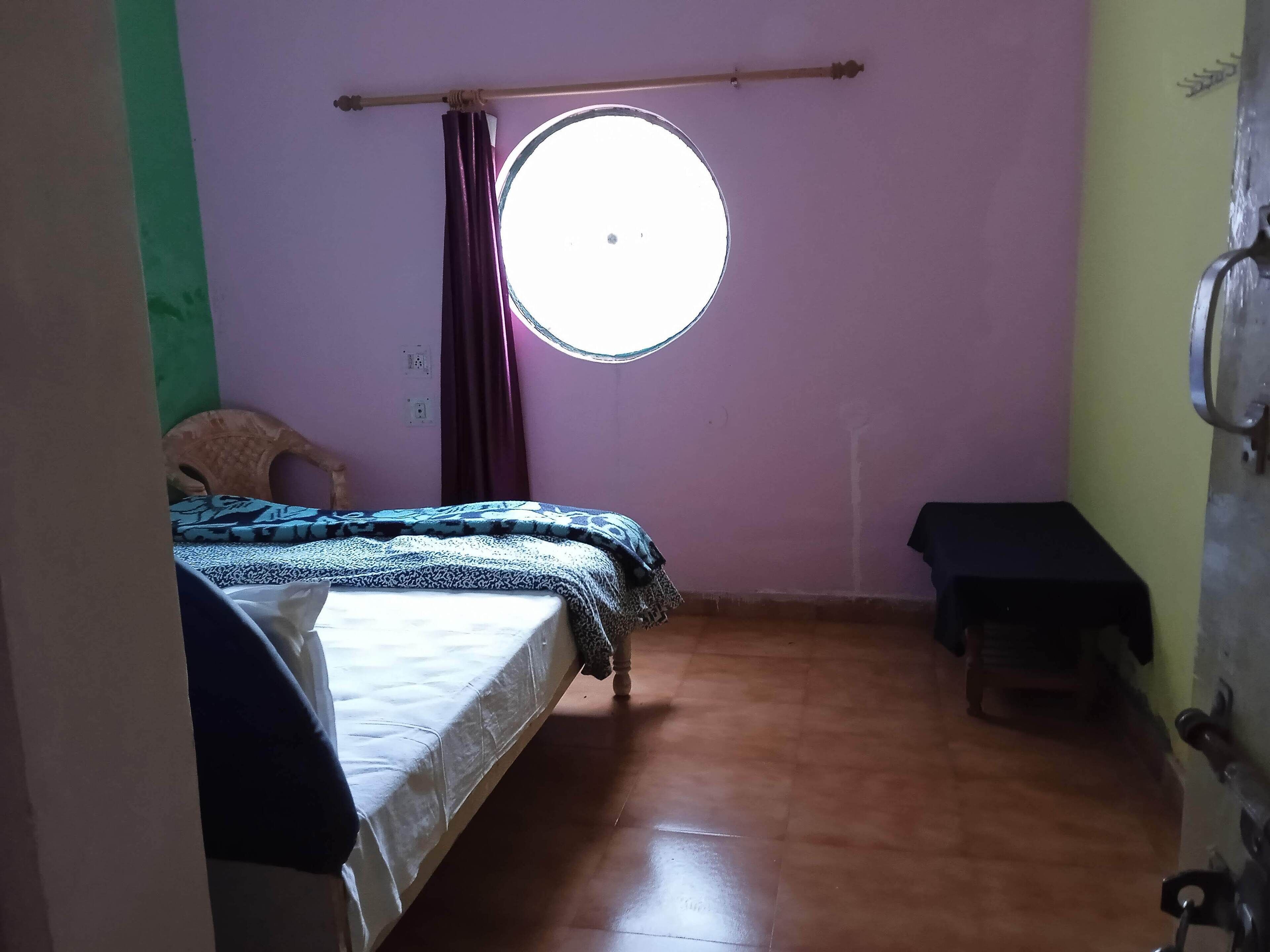 Una habitación con una cama, un taburete pequeño, una ventana y una puerta.