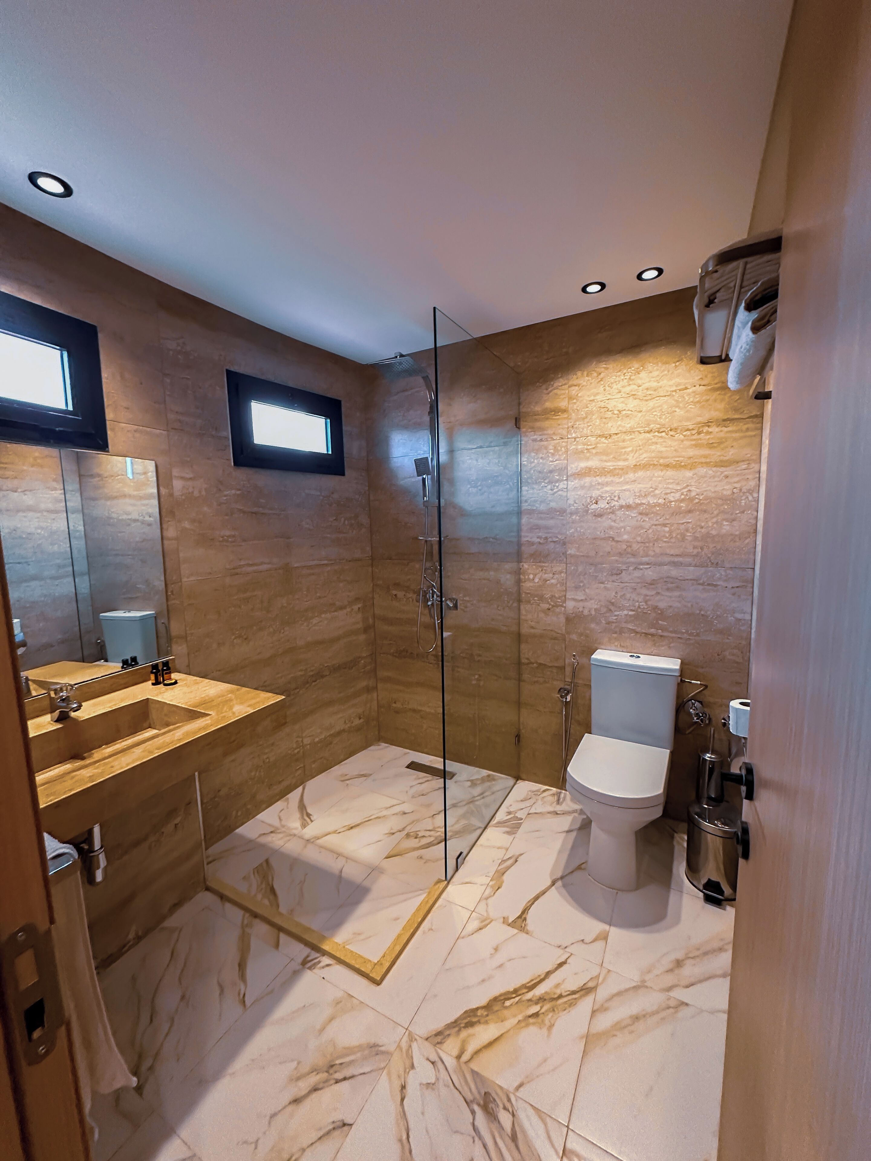 Et moderne badeværelse med en glasafskærmet brusekabine, et marmorbelagt gulv, et træbord og et toilet.