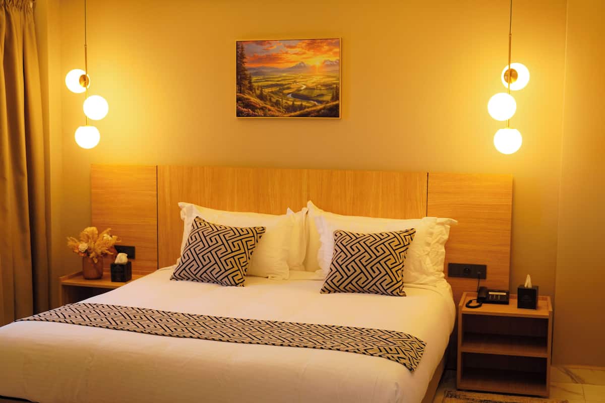 A neatly made bed with patterned pillows, a bedside table with a vase of flowers, and a painting above the bed.