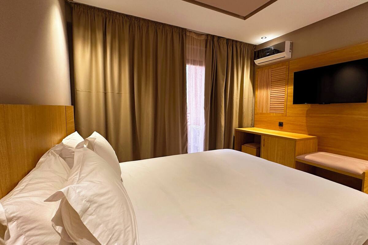 A hotel room with a large bed, a wooden headboard, a flat-screen TV mounted on the wall, a small desk, and a bench.