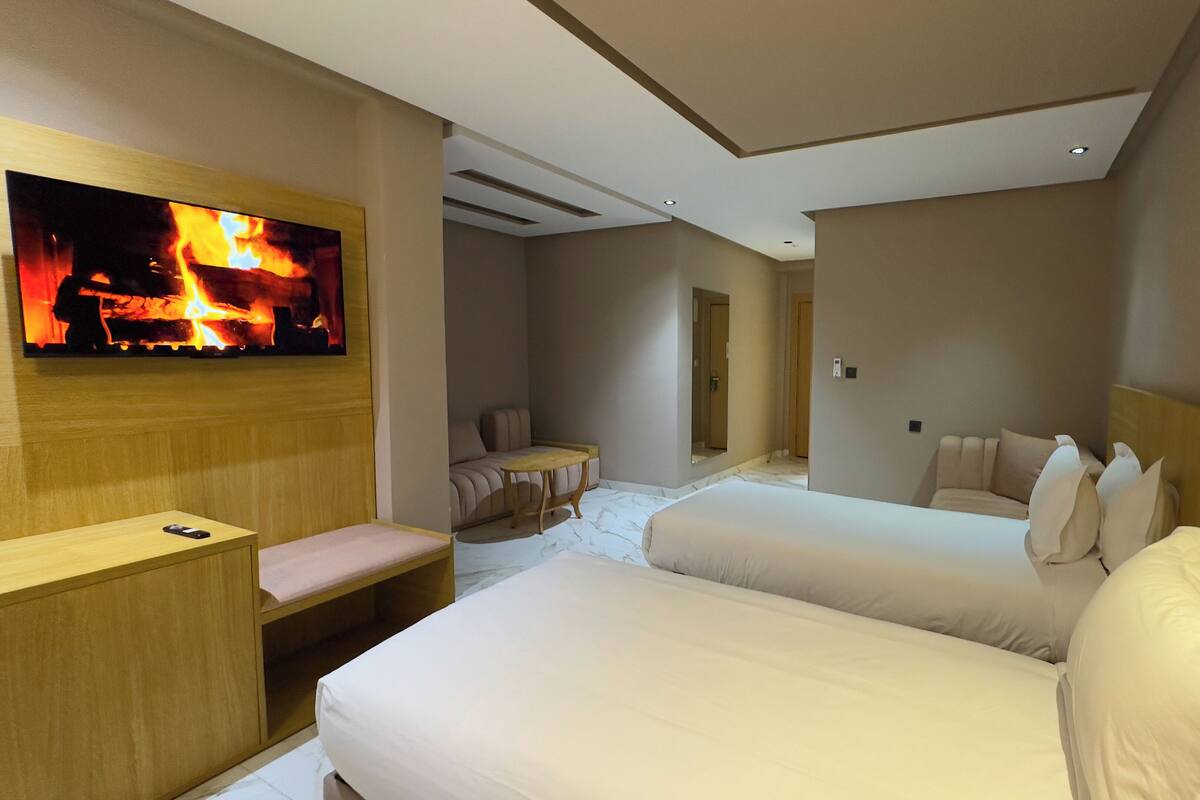 A hotel room with two beds, a TV displaying a fireplace, a small table, and a bench.