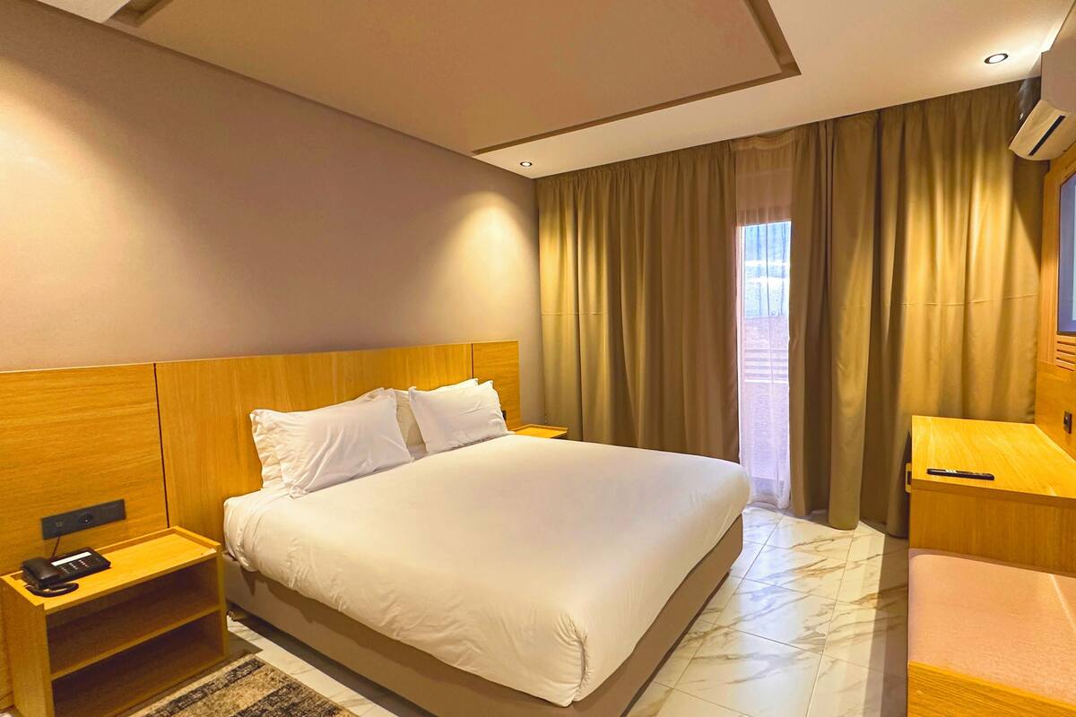A hotel room with a large bed, a bedside table with a lamp, a TV mounted on the wall, and a window with curtains.