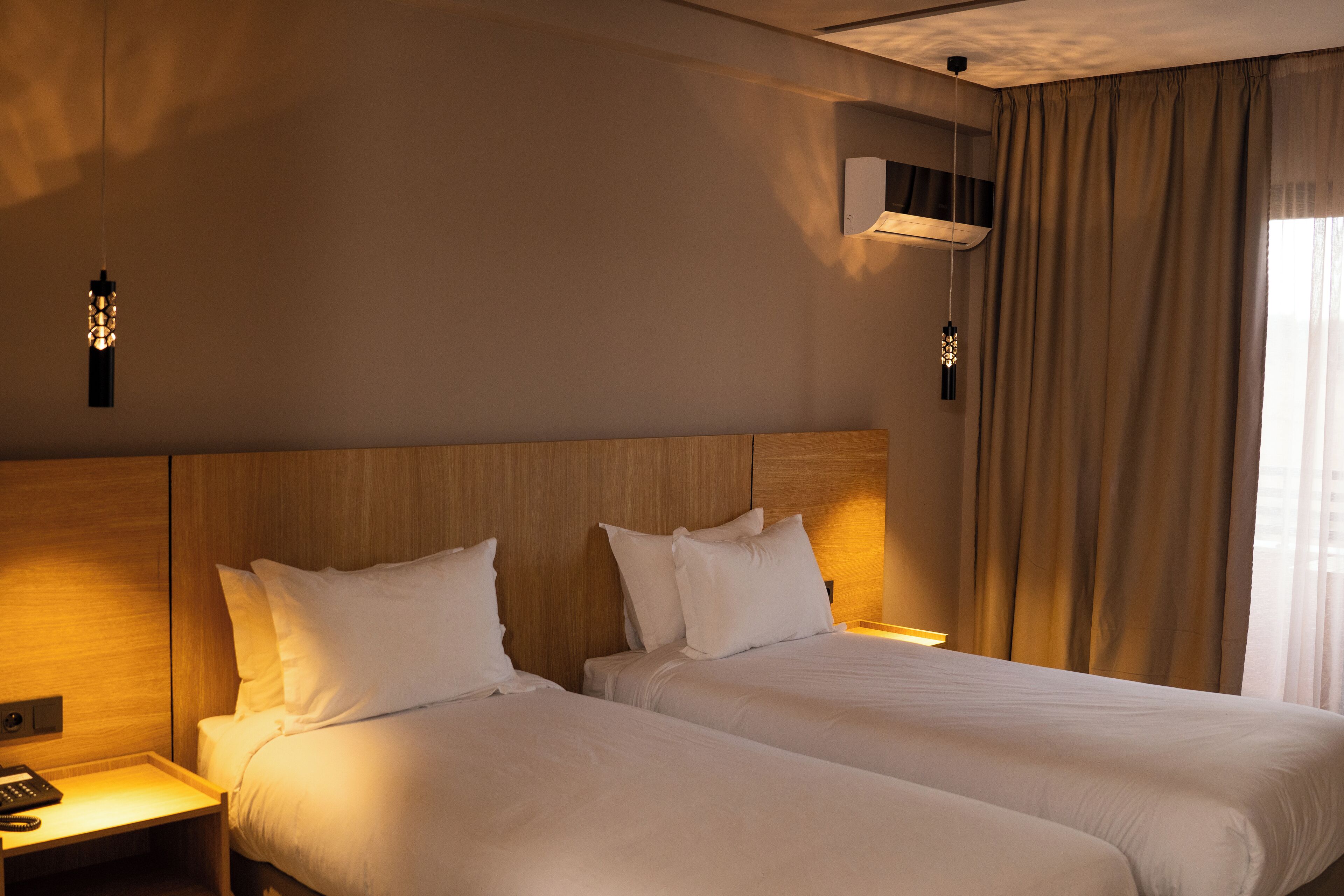 Ein Hotelzimmer mit zwei Betten, einem Holzkopfstück, Nachttischlampen und einem Fenster mit Vorhängen.