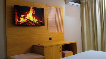 A hotel room with a TV mounted on a wooden wall, a bed, a wooden desk with a chair, and a window with curtains.