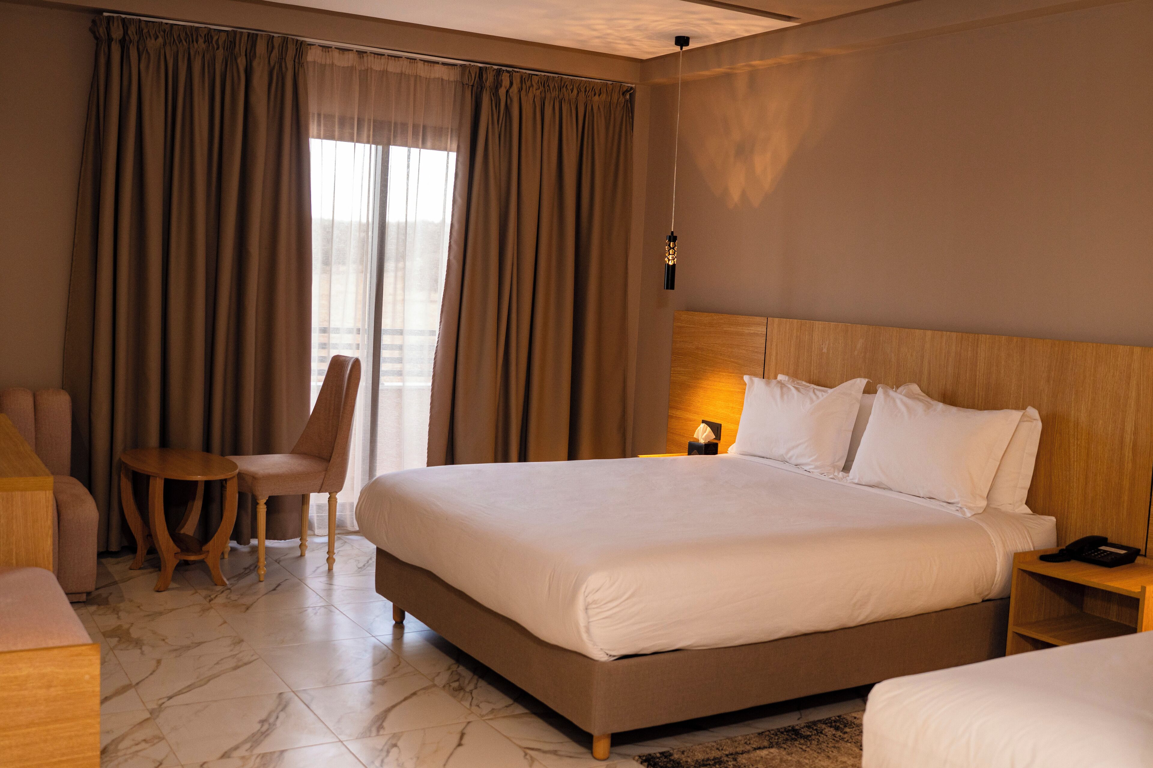 A hotel room with a large bed, a chair, a small table, and a nightstand with a lamp.