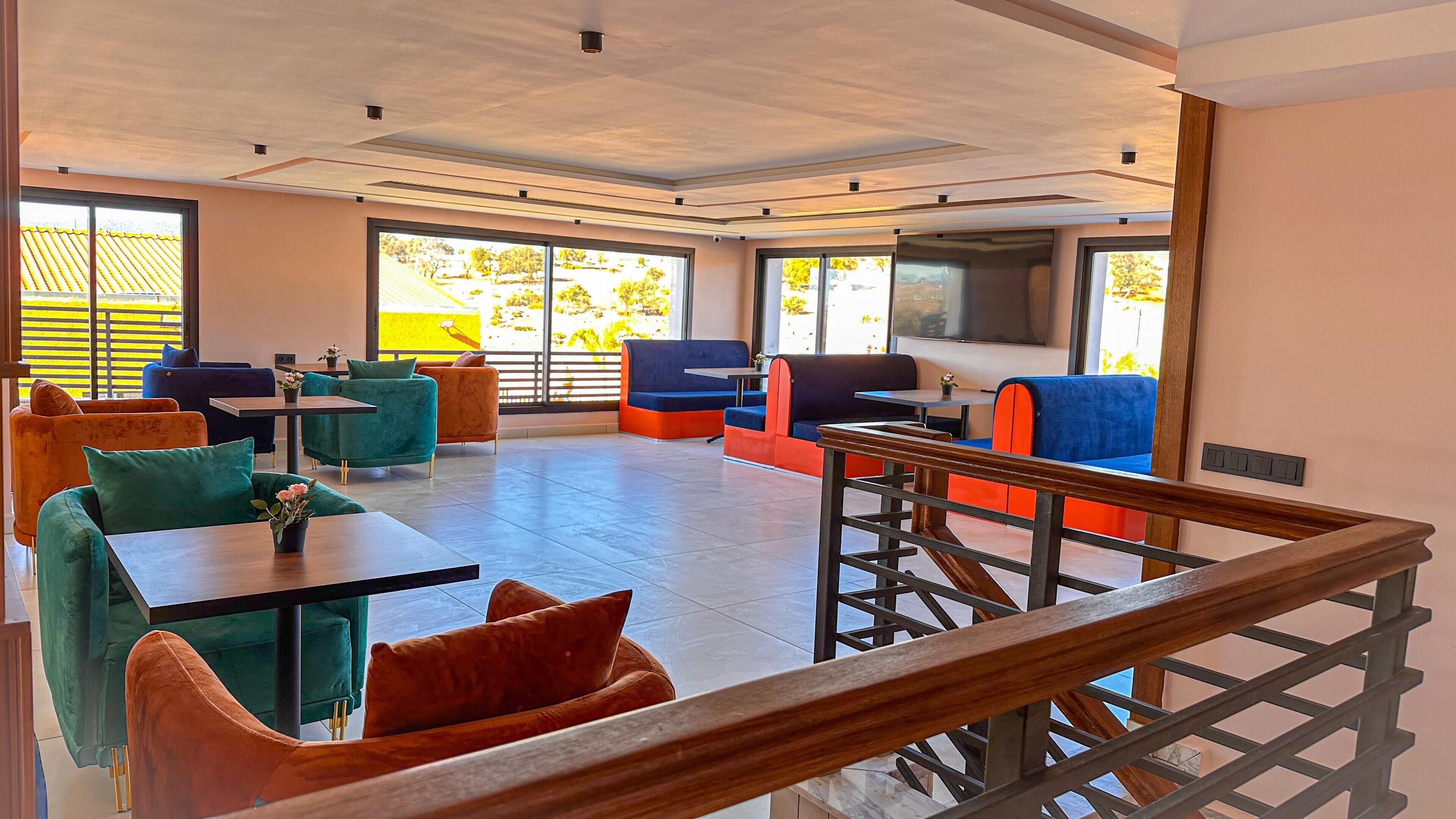 Un espace intérieur spacieux avec de nombreuses tables et chaises, de grandes fenêtres donnant sur un paysage verdoyant et une rambarde en bois.