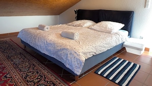 Une chambre avec un plafond en pente, un lit recouvert d’une literie à motifs floraux, deux serviettes enroulées, une table de chevet, une prise de courant, un tapis à motifs et un tapis rayé.