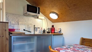 Une cuisine compacte équipée d’un four à micro-ondes, d’un évier et d’un petit réfrigérateur. On y trouve une table en bois recouverte d’une nappe à motifs floraux, ainsi qu’une chaise.