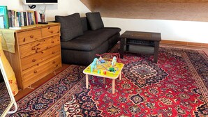 Un salon chaleureux avec un canapé gris foncé, une commode en bois et une petite table avec des jouets. Un grand téléviseur à écran plat est fixé au mur, et un tapis à motifs recouvre le sol.