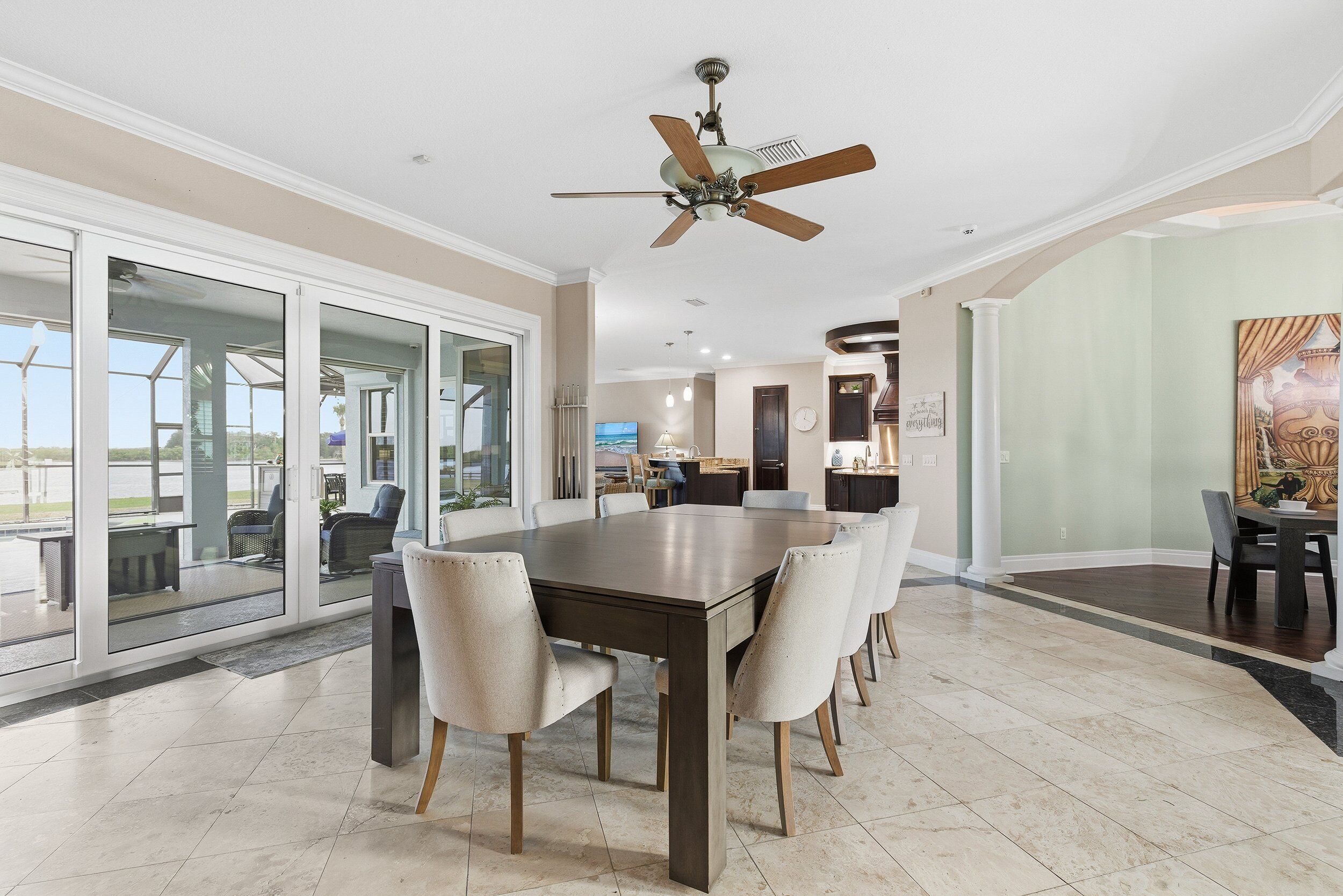 A spacious dining area with a large table and chairs, a ceiling fan, and large windows offering a view of the outdoors.