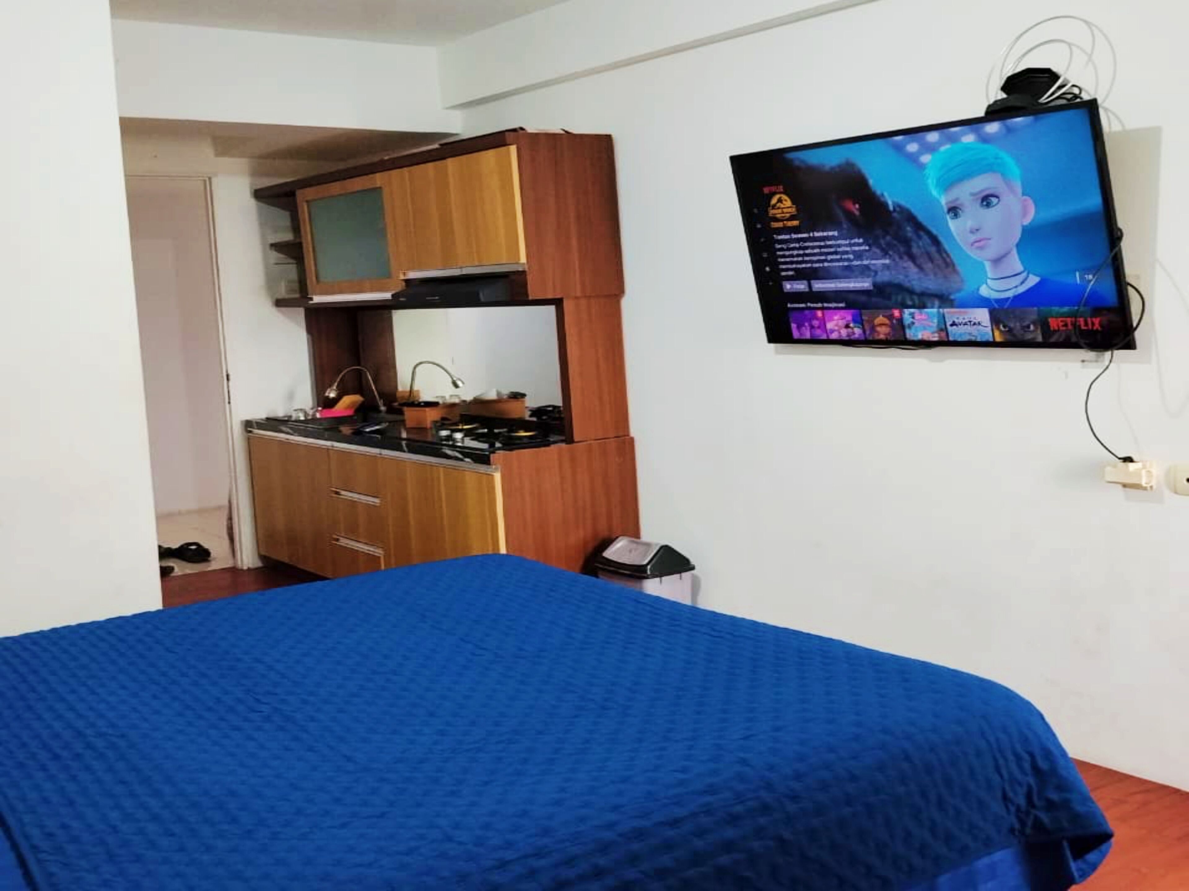 A bedroom with a bed, a TV mounted on the wall, a kitchenette, and a door leading to another room.