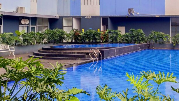 Una piscina con borde de azulejos azules, rodeada de vegetación y un edificio de varios pisos con balcones.