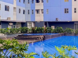 A swimming pool with a blue tiled deck, surrounded by greenery and a multi-story building with balconies.