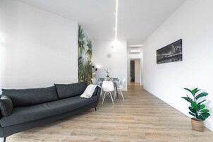 A modern living room with a dark grey sofa, a dining table with chairs, a potted plant, and a cityscape painting on the wall.
