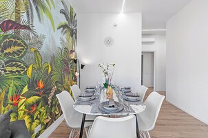 A dining area with a tropical mural, white chairs, and a table set for six with plates, glasses, and a vase of flowers.