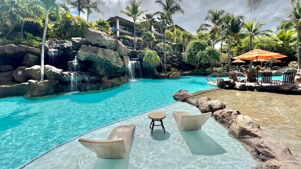 Une piscine aux eaux cristallines, agrémentée d’une cascade, entourée de rochers et de palmiers, avec des chaises longues et des parasols.