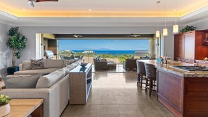 Un vaste espace de vie comprenant un grand canapé, une table à manger et un îlot de cuisine. Une porte ouverte offre une vue sur l’océan.