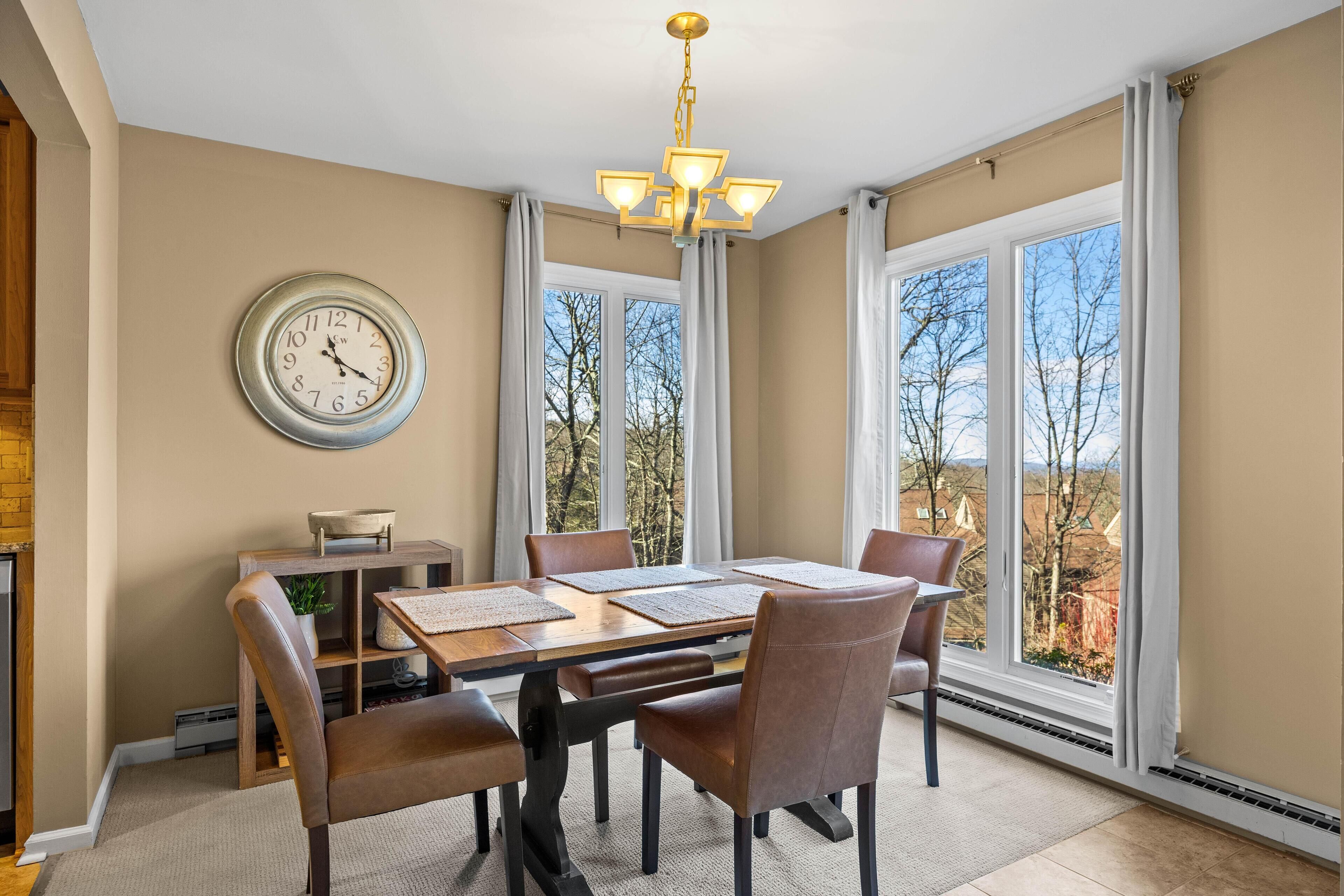 A dining room with a large table, chairs, a clock, and a view of trees through large windows.