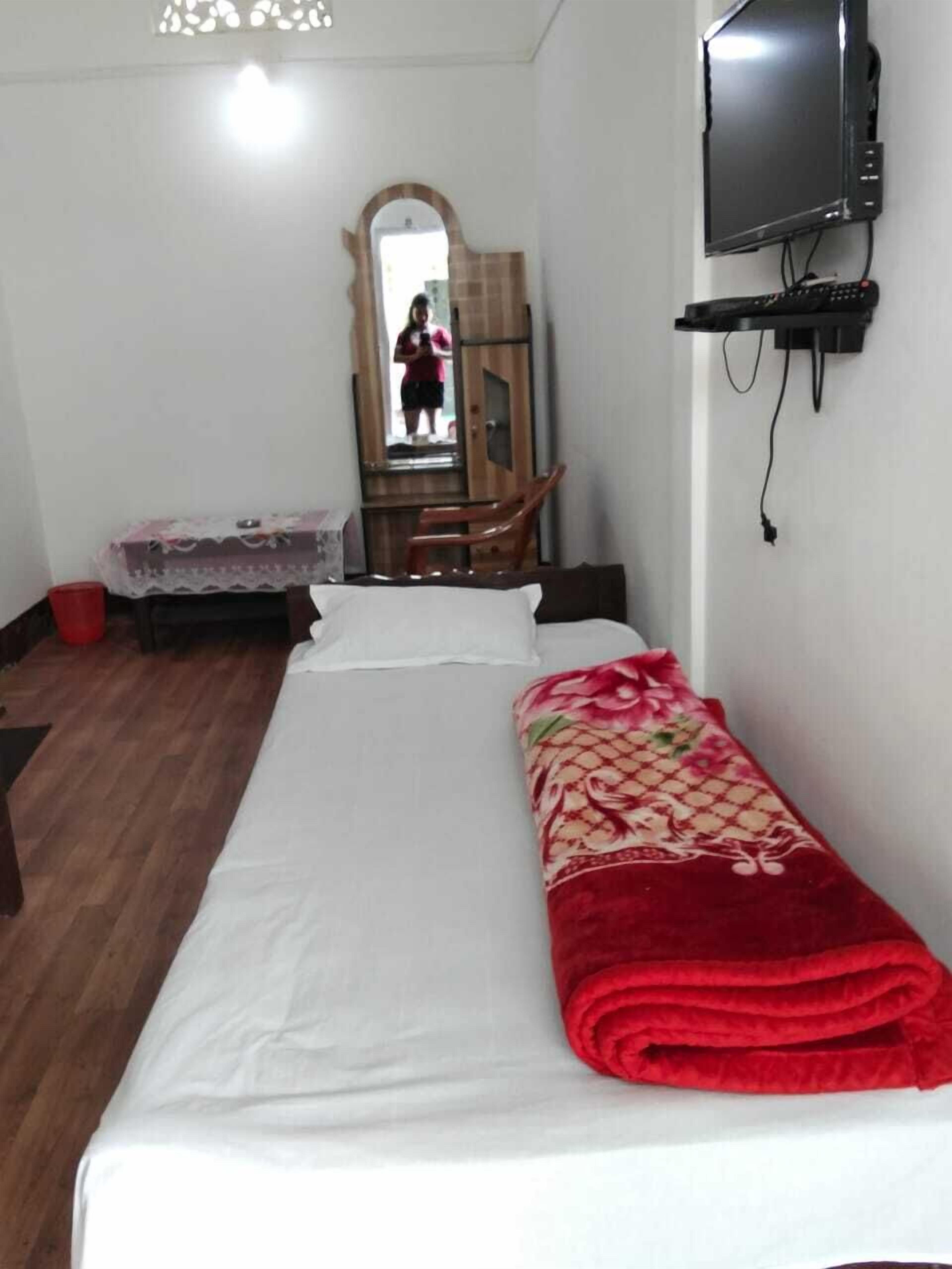 Een enkel bed met een wit laken en rode deken, een houten stoel, een klein tafeltje met kantkleed, een aan de muur bevestigde televisie en een spiegel met houten lijst.