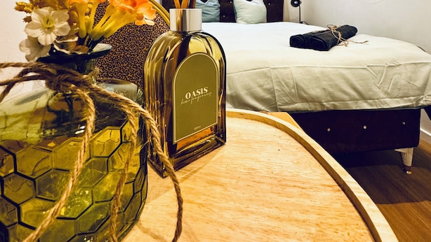 Ein Hotelzimmer mit einem Bett, einem Nachttisch mit einer Oasis Körperparfumflasche und einer Vase mit Blumen.