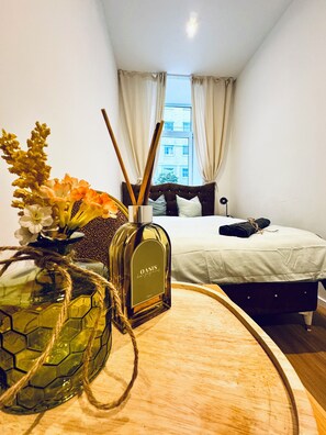 A hotel room with a bed, a bedside table with a bottle of Oasis body perfume, and a vase with flowers. - Cozy Apartment Near Matzleinsdorfer (Vienna)