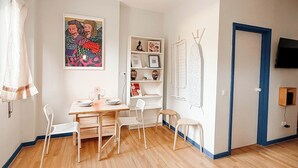 A room with a wooden floor, a dining table with chairs, a bookshelf, a framed picture, a door, and a television mounted on the wall. - Cozy studio in Porto (Porto)