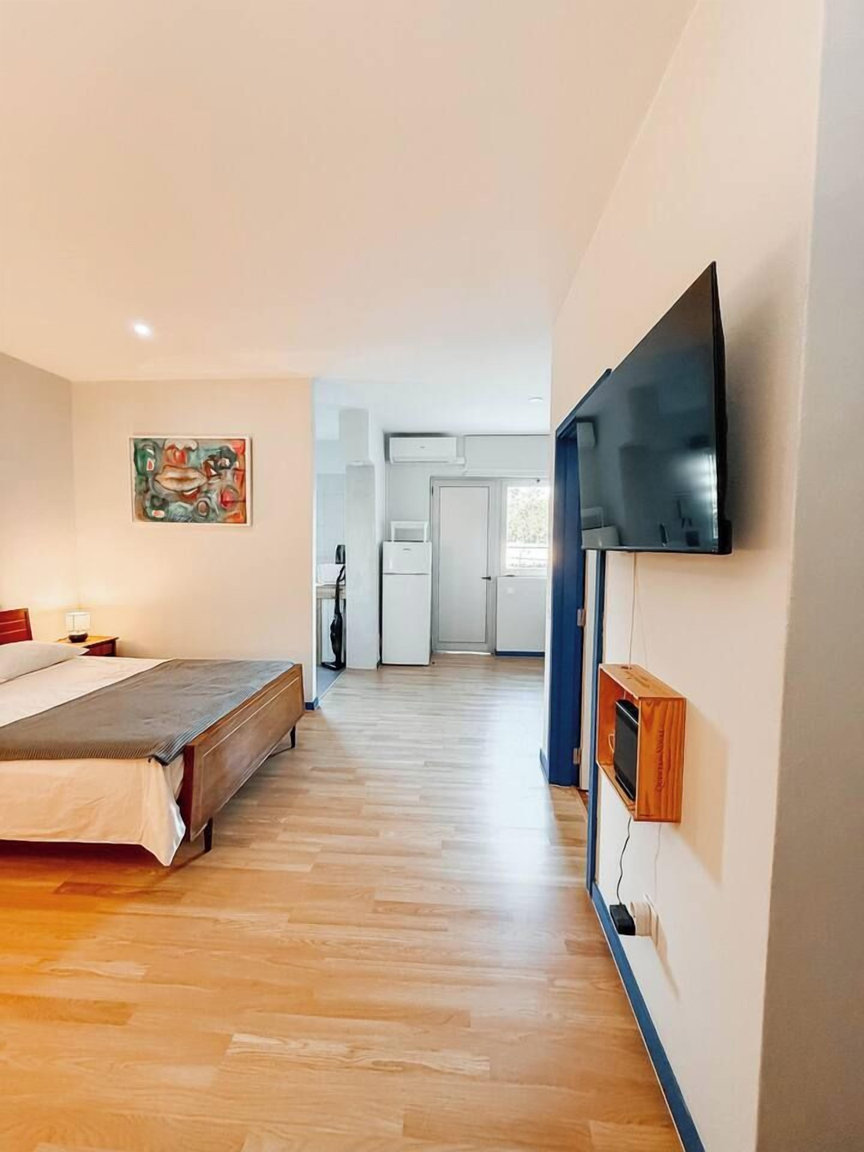 Ein Schlafzimmer mit einem Bett, einem an der Wand befestigten Fernseher und einer Küchenzeile im Hintergrund.