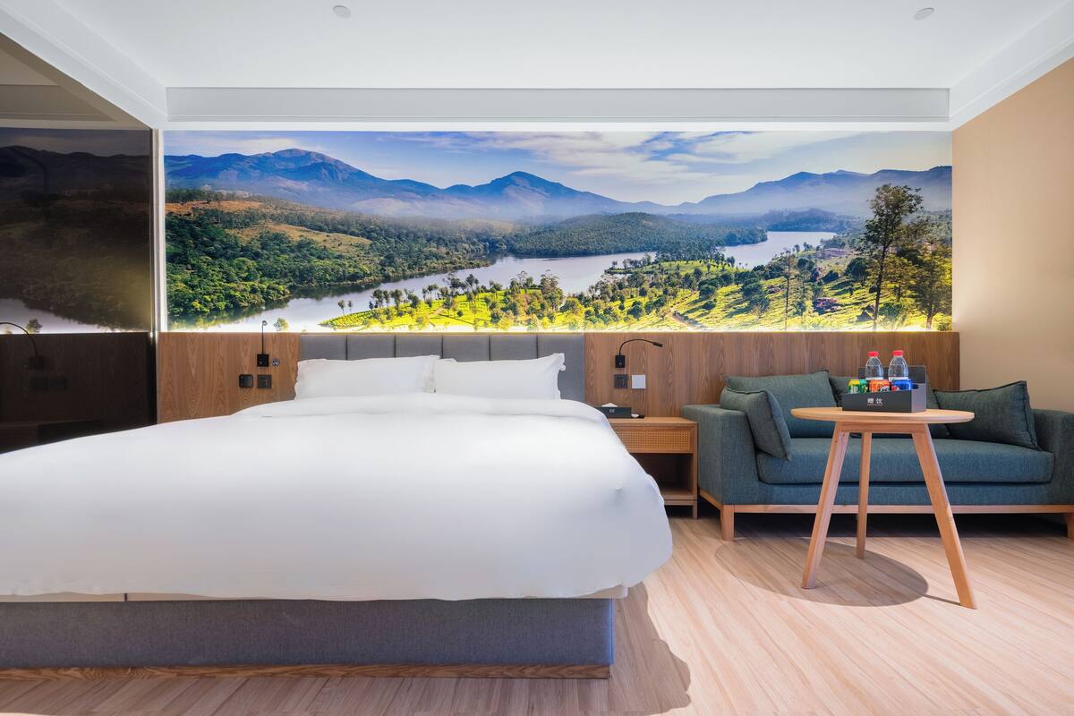 A modern hotel room with a large bed, a green sofa, a small wooden table, and a mural of a mountainous landscape.