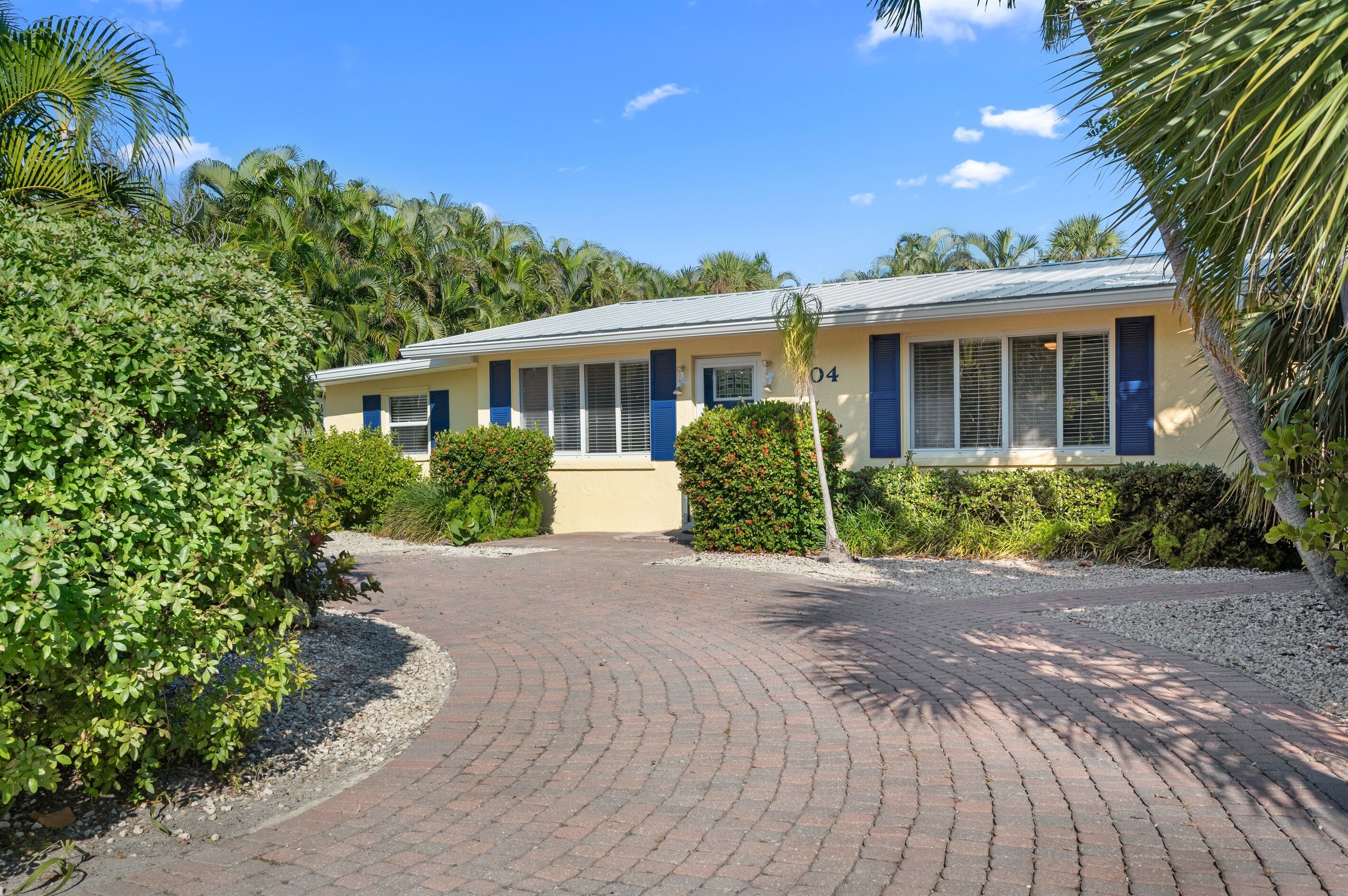 A building with a paved driveway leading to a gated entrance, surrounded by lush greenery and palm trees.