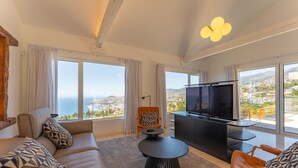 Un salon moderne avec un canapé, une table basse ronde, un téléviseur et de grandes fenêtres donnant sur la mer.