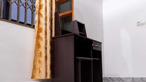 Ein Zimmer mit einem dunklen Holzschrank, einer weißen Wand, einem Fenster mit Metallgitter und einem gemusterten Vorhang.