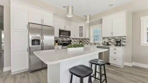 Una cocina moderna con armarios blancos, electrodomésticos de acero inoxidable, una isla central con taburetes e iluminación colgante.