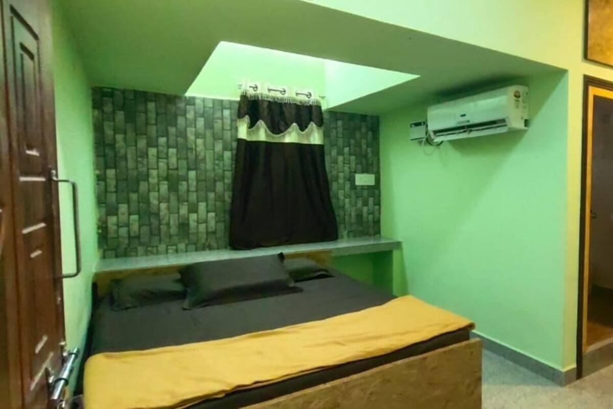 A small, compact room with a bed, a window with curtains, an air conditioning unit, and a door leading to another room.