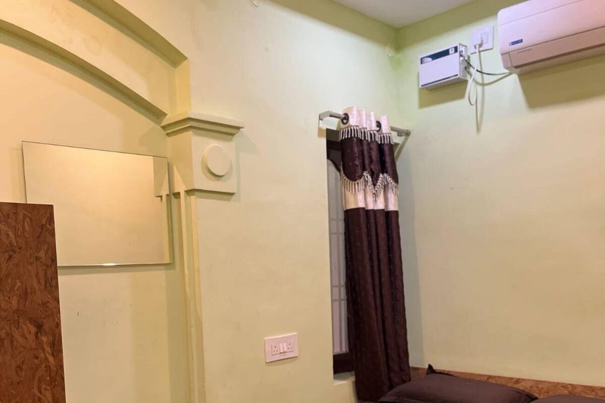 A room with a bed, a wooden bench, a window with curtains, an air conditioner, and a ceiling fan.