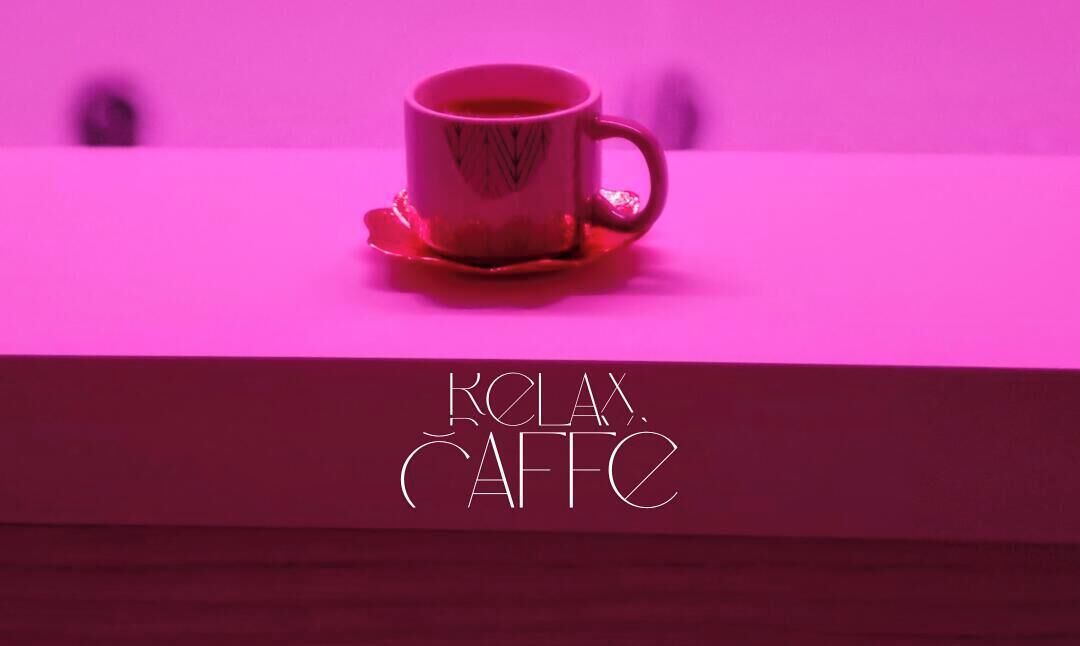 A cup of coffee on a saucer with a pink background and the text 'KOLA CAFE' on a wooden surface.