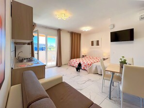 A hotel room with a kitchenette, a bed with a floral bedspread, a TV mounted on the wall, and a balcony with a view of buildings. - Bright Studio Close to the sea (San Michele al Tagliamento)