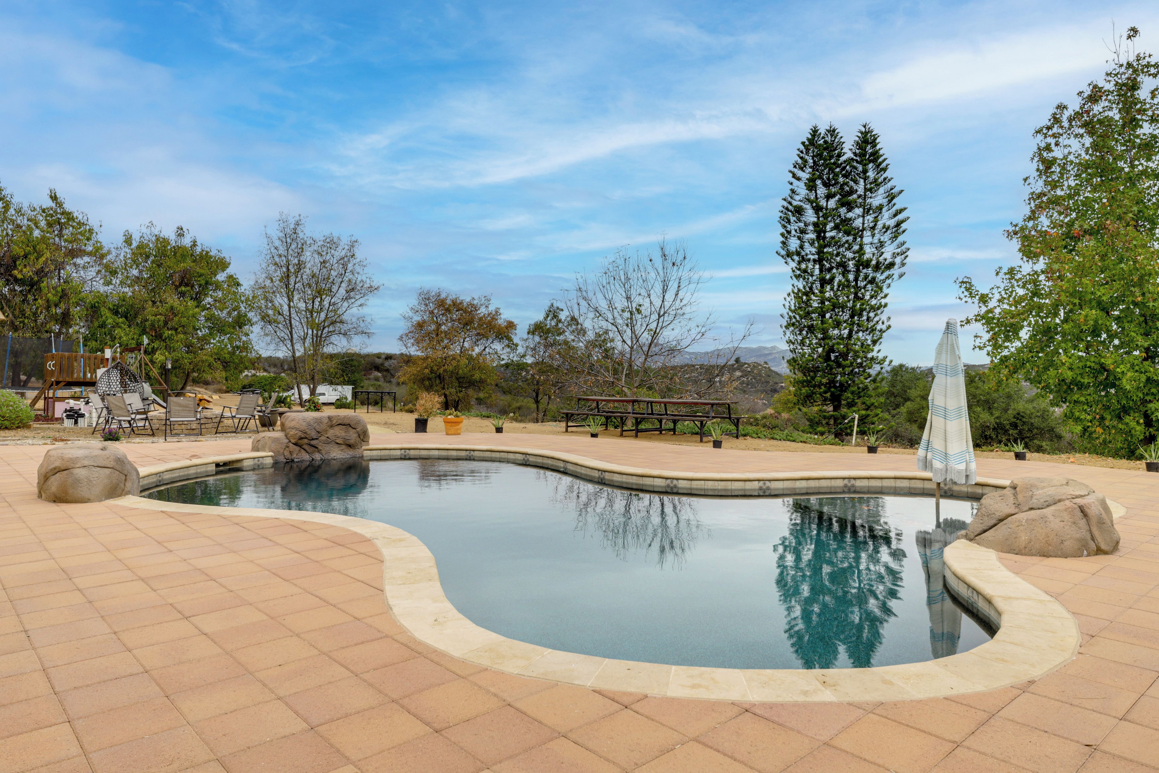Une piscine à bord incurvé, entourée d’une terrasse carrelée. On aperçoit au fond des chaises longues et une table de pique-nique.