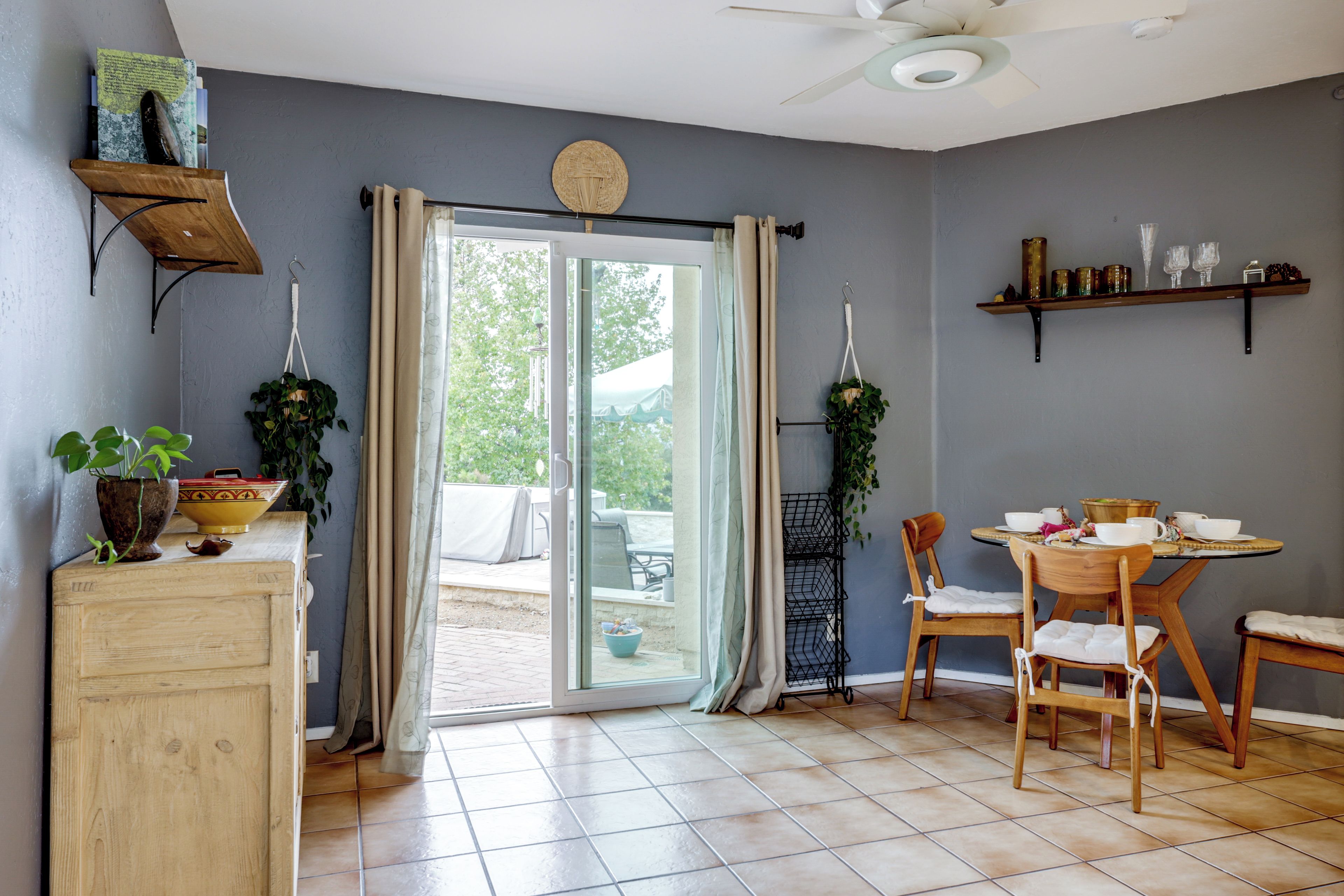 A room with a sliding glass door leading to an outdoor area, a wooden dining table with chairs, a ceiling fan, and shelves with decorative items.