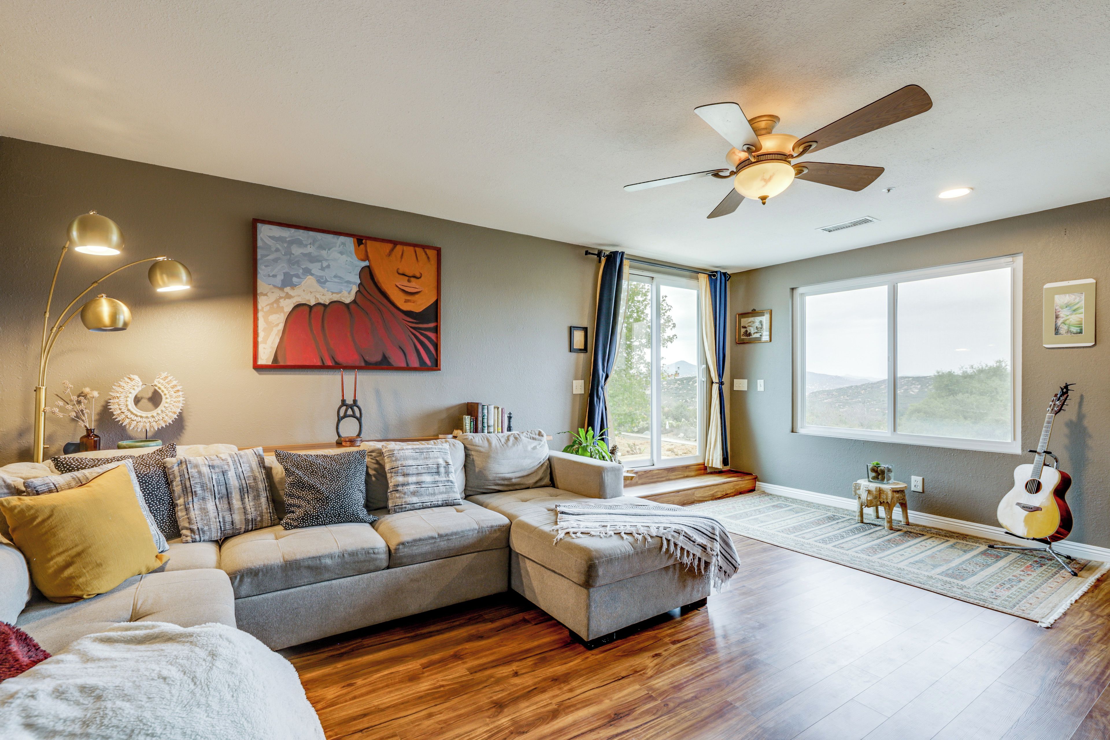 A living room with a sectional sofa, a ceiling fan, a guitar, and a large window.