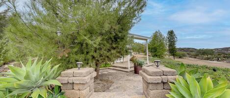 A stone archway leading to a covered area with a pergola, surrounded by large green plants and a clear sky.