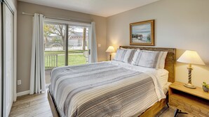A bedroom with a large bed, a nightstand with a lamp, a painting on the wall, and a view of the outside. - Resort Perks & Family Fun! St George Retreat (St. George)