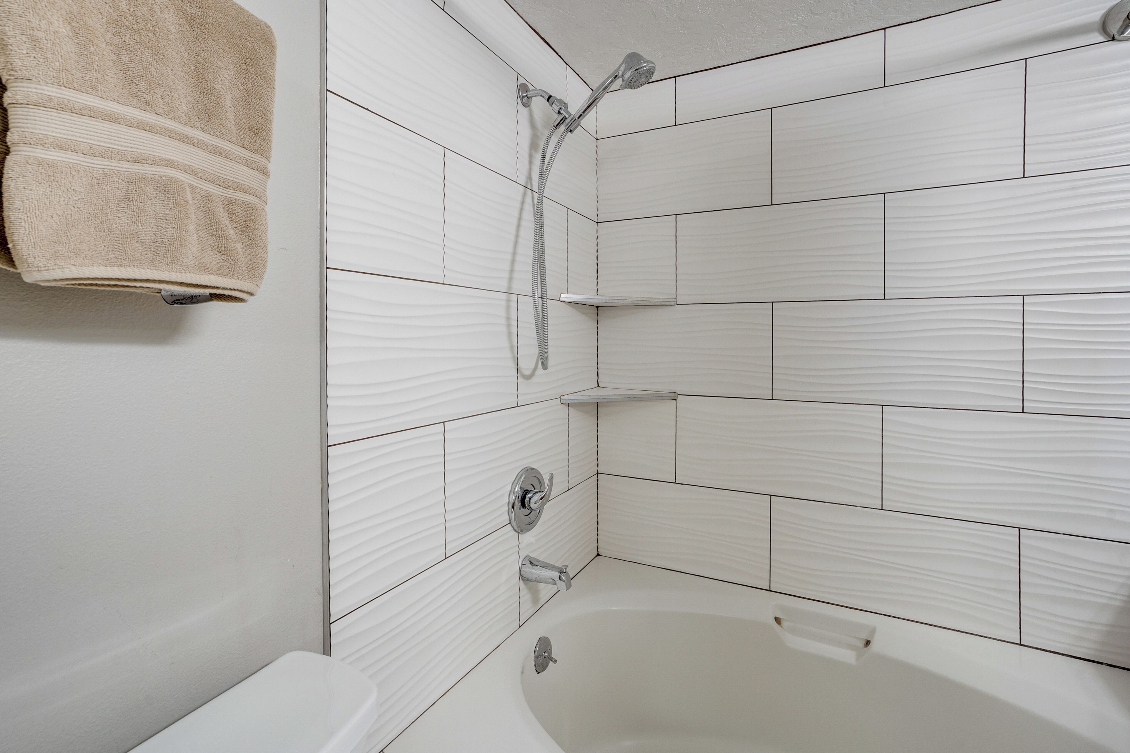 Een badkamer met witte betegelde wanden, een bad, een douche met een handdouche, en een handdoek aan de muur.