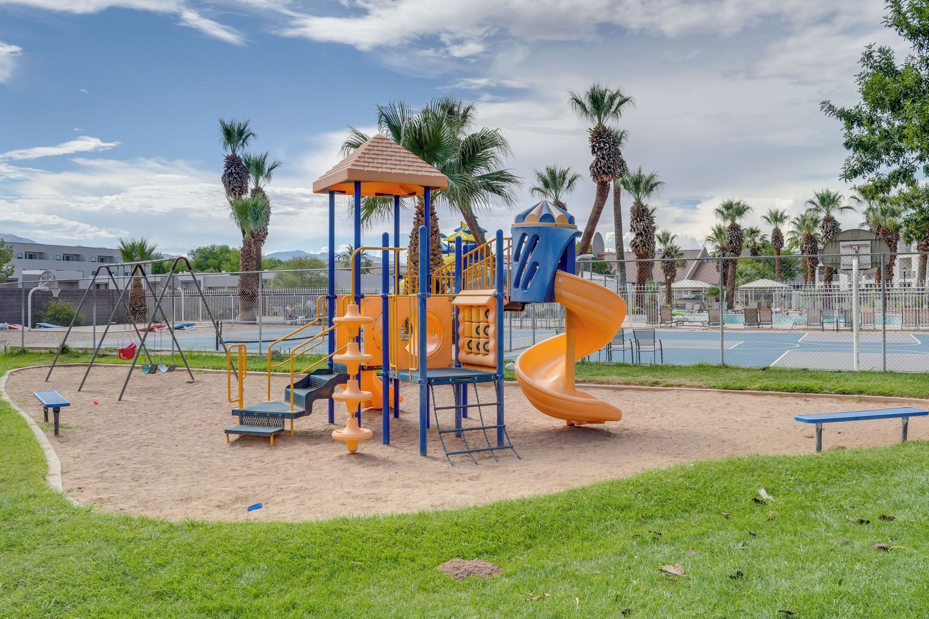 A playground with a slide and climbing structures, surrounded by palm trees and a fence.